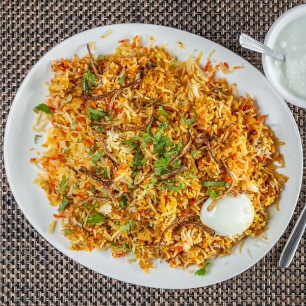 Indian biryani with rice, spices, cilantro, fried onions, and a hard-boiled egg on a white plate.