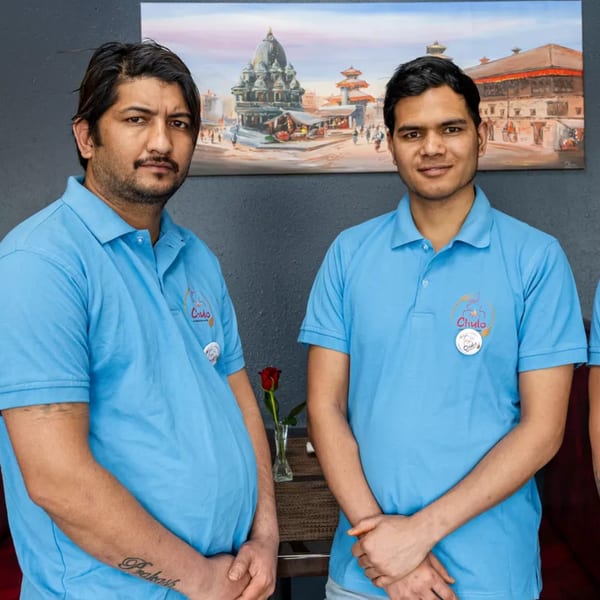 Two men in blue Chulo Cuisine of Nepal & India shirts stand before a painting of a Nepalese temple.