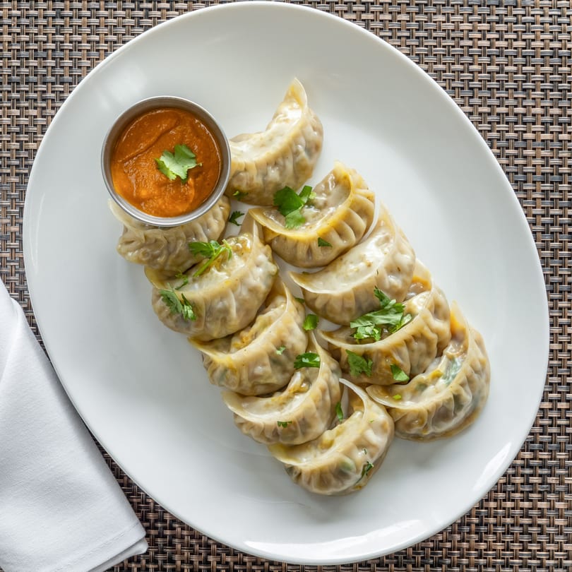 A white plate of Chulo Cuisine momos, garnished with cilantro, and a side of orange dipping sauce.