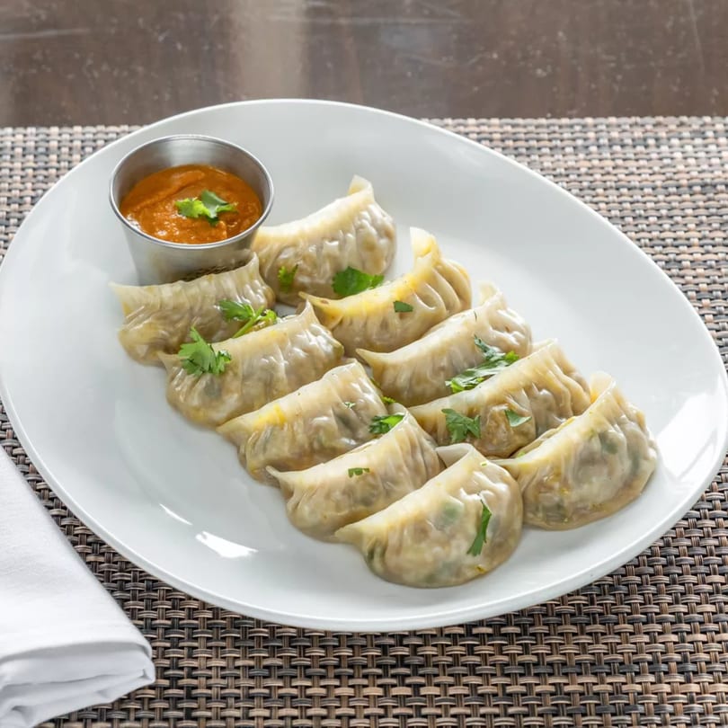 A white oval plate holds twelve steamed momos, garnished with cilantro, and a side of orange dipping sauce.