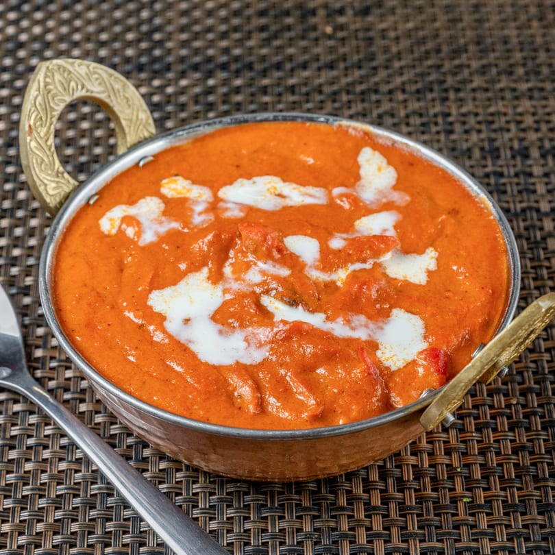 Indian butter chicken, a creamy orange curry with white swirls, in a decorative copper bowl.